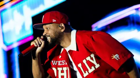 EPA Will Smith sings into a red hand-held microphone. He is wearing a red cap and a red shirt which is open. 'West Philly' is written across the shirt in white. He is on a brightly-lit stage.