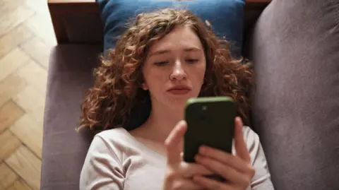 Getty A teenager lies on a grey sofa looking at her phone, which has a green case. She has curly red hair and freckles, and wears a cream top.