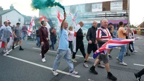 PA Media Protesters walking along a road wearing and carrying union jacks and St George cross flags with the man in the centre of the picture carrying a green smoke grenade