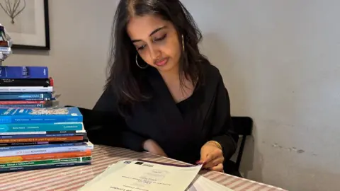 BBC A young woman with long black hair, wearing a black top and gold hoop earrings. She is sitting at a table and looking at a pile of exam certificates. There's a stack of textbooks next to her, and a plain white wall behind her.