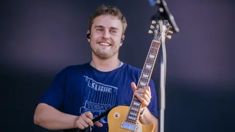 Thomas Niedermueller/Getty Images Sam Fender performing onstage during Southside Festival at Take-off Gewerbepark on 22 June, 2025 in Neuhausen, Germany.  He has short, curly brown hair and stubble on his face.  He is smiling and holding up a yellow electric guitar.  He is weaing a navy t-shirt with a "Classic Today" logo and has a set of black earbuds in his ears. 