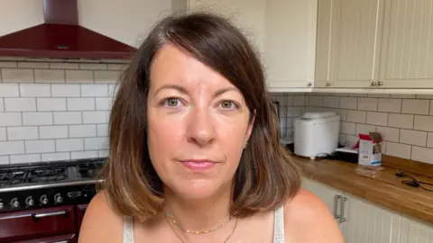 BBC A woman with a brown bob. She is looking at the camera and is sitting in a kitchen, with a red stove in the background
