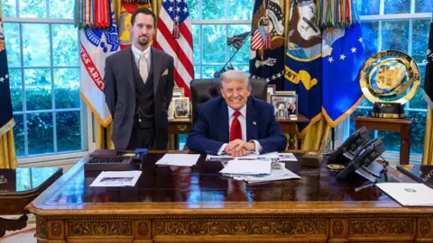 White House Trump sits behind desk in blue suit and red tie while Anotni stands in three-piece gray suit with a beige tie, white collar and watch chain in front of windows covered in colorful flags and banners