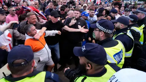 PA A line of police officers on the right of the picture are faced with a group of protesters, several of who are carrying flags and signs. One man stands holding his phone towards the confrontation