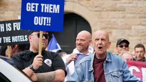 PA Media Two men at a protest - one wears a black t-shirt and sunglasses and is holding a sign which reads "get them out!!". The other man is balding, wears a denim jacket and purple t-shirt, and appears to be shouting