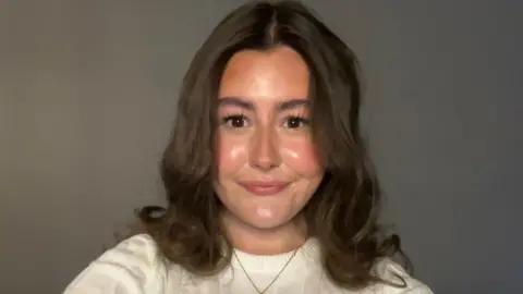 Aoife Aoife, a 16-year-old girl, taking a selfie. She is wearing a white knitted top and has brown, curly hair that goes to her shoulders. She is wearing a gold chain around her neck and is in front of a neutral background
