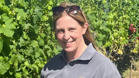 BBC Gwen, who has long blonde hair with sunglasses pushed up on her head, wearing a grey polo shirt and standing in a vineyard, with a row of plants behind her. 