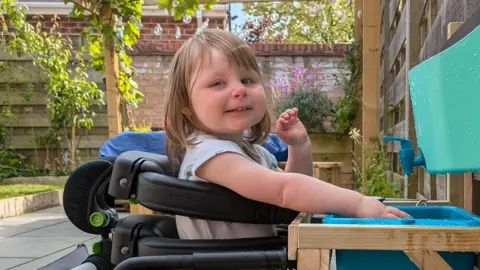 Gracie Davies A three-year-old girl in a wheelchair playing with water. She is smiling at the camera. She has long brown hair and a fringe. She is in a garden on a sunny day.