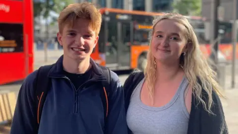 BBC Students Evan Stoddart and Holly Wells, who stand in front of Wood Street near Central Square, Cardiff. Evan (left) has ginger short hair, and wears a dark blue quarter zip fleece and a backpack, while Holly (right) has blonde long hair and wears a grey vest and cardigan. There are buses in the background, which are blurred as the two people, in the foreground are the focus of the picture.