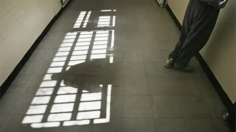 Getty Images A corridor with sunlight streaming through a barred window, casting shadows on the tiled floor. A person stands against the right wall, only their lower body visible. Their shadow on the floor.