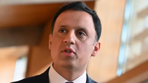 Getty Images Anas Sarwar, a man with black hair, looking to his right in the Scottish Parliament. He is wearing a dark suit and white shirt in a close-up shot. There is wood panelling behind him that is out of focus. 