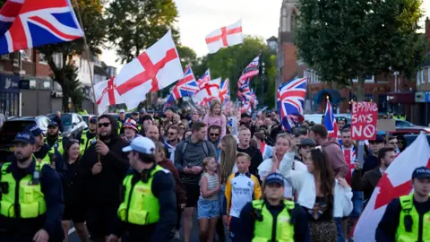 PA Protesters marching in a tight group, marshalled by a line of police officers at the front. The protesters are waving England and union jack flags. There are a handful of women and children near the front of the crowd.