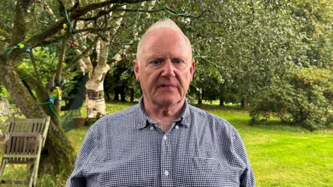 A man with short grey hair looks into the camera. He wears a navy and white chequered shirt. Behind him a plot of green grass and several trees with green leaves.