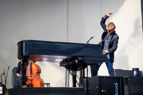 Getty Images Tom Odell jumps up from his piano, with a fist raised to the air, during a festival performance in 2025