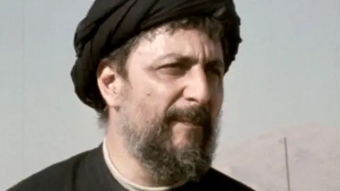 Imam Sadr Foundation A black and white image of Musa al-Sadr. He has a beard, is wearing a hat and is looking thoughtful. 
