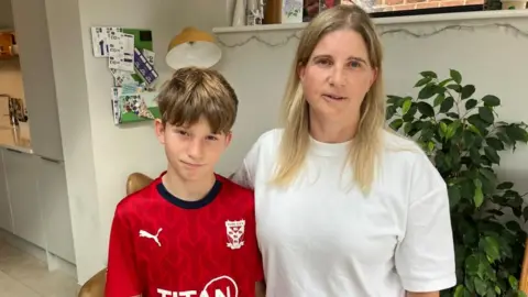 BBC/Seb Cheer A boy and his mum look into the camera, in a room with white walls and a plant behind them. There is also a noticeboard with coupons on it, and fairy lights. He is wearing a York City FC football shirt, and she is wearing a white T-shirt.