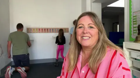 Louise Shore sits and smiles for the camera. She is wearing a large pink short with red stripes and has long straight blond hair. In the background a man and child can be seen painting a while wall.