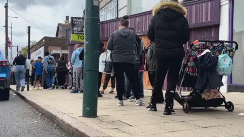 A queue has formed outside a uniform shop. People are standing and waiting. Many are wearing jackets and carrying bags. One woman has a pushchair. A car is parked in the road. Other businesses can be seen in the street. There is a To Let sign on one property.