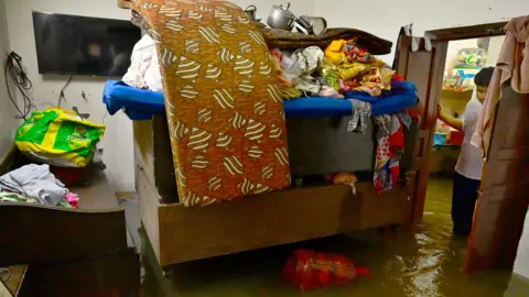 Hindustan Times via Getty Images Sofa, bed and other furniture submerged in water due to floods as a man stands at the gate and stares at his losses in Punjab, India.