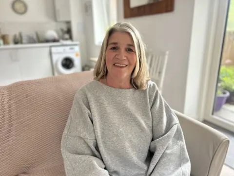 Claire, who is wearing a grey jumper and has blond straight air, sits on a sofa in a room with the kitchen visible behind her