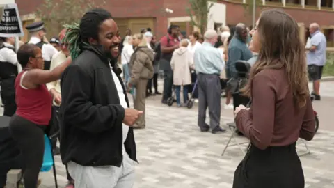 JJ standing among a group of people. He is wearing a black jacket over a white top and grey trousers