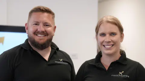 A man and a woman in matching black polo shirts that say Crossroads on them. The main has short brown hair and a brown beard and the woman has blue eyes and strawberry blonde hair in a ponytail