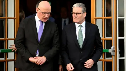 PA Media John Swinney, who is bald with glasses and Sir Keir Starmer, grey-haired with glasses, walk through open pane glass doors. Swinney is on the left and Starmer is on the right. Swinney, with his head slightly bowed, is speaking. Both are wearing black suits and white shirts. Swinney is wearing a purple tie, Sir Keir a green tie.
