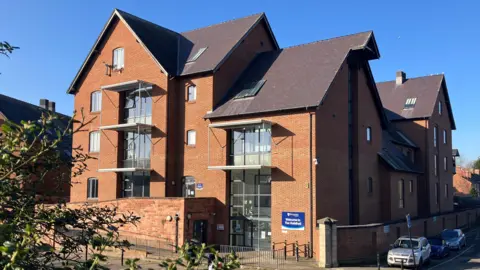 A large red-bricked building with various gabled roofs. A sign outside a glass door on the ground floor reads "welcome to the Guildhall". Three cars are parked along the side of the building.
