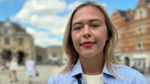 Daisy Blakemore-Creedon wearing a blue and white striped-shirt over a white T-shirt smiles at the camera as she is photographed outside a building.