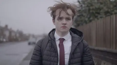 Judah Cousin/More Than What You Are A still from Judah Cousin's film More Than What You Are. He is walking down the street wearing his school uniform. He has a sad look on his face. He has a black puffa coat on, white shirt and red tie. His ginger hair flies in the wind.