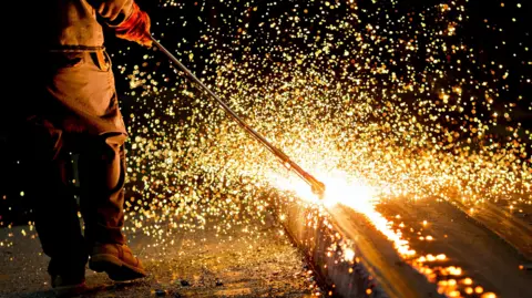 Bloomberg via Getty Images A steelworker runs a long metal tool along a steel plate causing countless sparks to fly in all directions. 