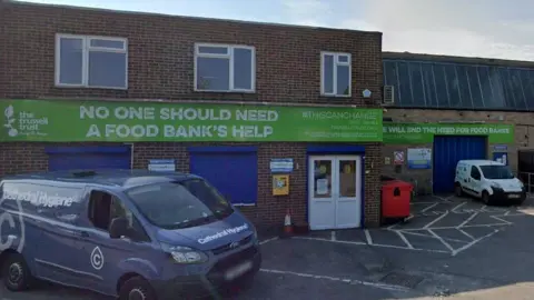 Google A view of a single-storey small warehouse in Salisbury, red brick. On it are big signs in green for the foodbank with the Trussell Trust logo. It says "No one should need a food bank's help".