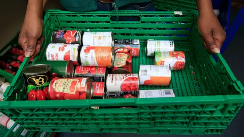 PA Media A shot looking down on a plastic green crate full of various brands of tinned tomatoes being held by someone, though you can only see their hands.