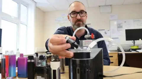 University of Bath/PA A man with close cropped dark hair and a grey and dark beard dropping liquid from a small black bottle into a table-top device with tubes running out of it. He appears to be in an office and has a number of bottles of vape liquid lined up on a table to his right.