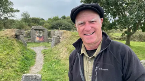 Archaeologist, Rhys Mwyn, is pictured next to graffiti on ancient stones and wears a black flat cap and fleece