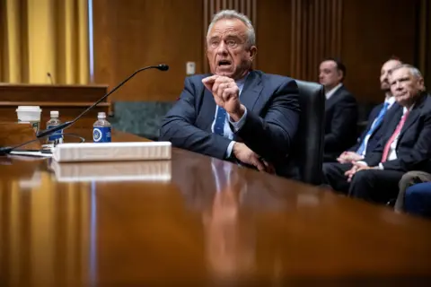 Politico AP Robert F Kennedy, sits at a wide wooden table. He is wearing a smart, tailored, dark suit. On the table is white folder, some plastic bottles and a white disposable drinks cup. He is speaking animatedly and gesturing with his left hand. In the background, other men in similar dark suits listen.