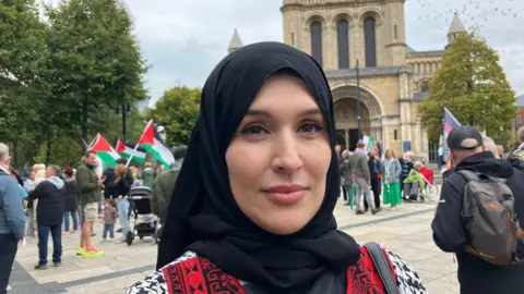Eftaima Najjair at the protest in Belfast, with St Anne's Cathedral in the background.  She is wearing a black hijab scarf on her her and a black and white pattered coat with red and black panels at the front.  Protesters holding Palestinian flags are standing in the distance behind her. 