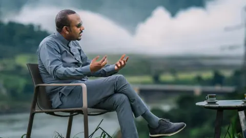 Office of Ethiopia's Prime Minister Abiy Ahmed in grey trousers and shirt sits on a chair with a coffee table in front of him. He is holding his hands out to make a point to an unseen interviewer. Behind him, blurred, are the gushing waters of the Blue Nile. 