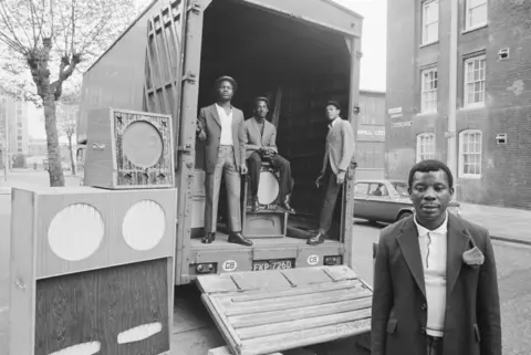 Dennis Morris Black and white photograph of three men standing in the back of a truck parked next to a sound system. A man is also stood in front of the truck.