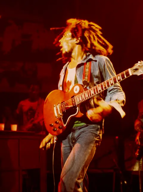 Dennis Morris Colour photograph of Bob Marley with a guitar slung across his body in a live performance.
