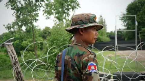 BBC/Lulu Luo A Cambodian officer stands guard next to a wall of razor wire. Beyond him are trees and a black screen.