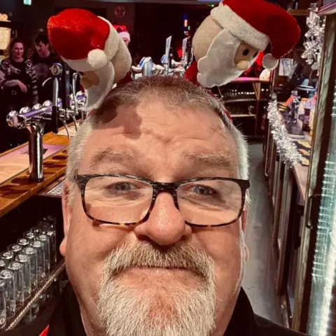 Graham Matthews A man with a white beard wearing glasses, he has a Christmas head band on with miniature Santa's. The picture is a selfie he's taken behind the bar of the pub.