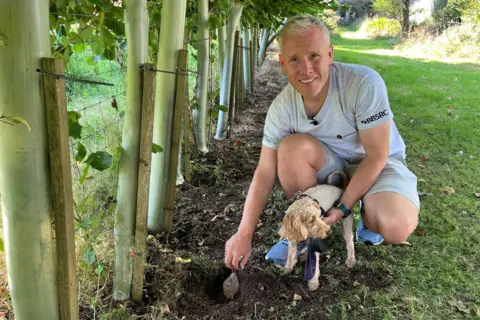 A man in a grey t shirt and shorts crouches below a row of trees while using a trowel to dig a hole. He's holding a small dog with curly hair wearing a collar.