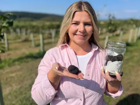 A woman with blonde hair and white t shirt with a pink shirt is holding a single truffle in her right hand and a jar of them in her left. She is standing in front of a junior orchard with blue skies.