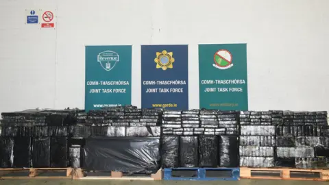 An Garda Siochana Four pallets have been lined up in a large warehouse. Each is packed with blocks of plastic wrapped packages shaped like bricks. Behind the drug haul are three signs each reading 'joint task force' in Irish and English. It also has a website address for Gardai