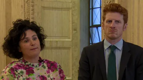 A wiry haired woman with blue eyes and flowery top and a ginger curly haired man wearing a navy suit, blue shirt and green tie looking concerned. They are standing in front of a yellow door frame.