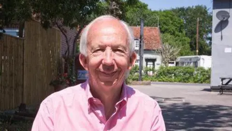 Neil Watts smiles at the camera while standing outside. He wears a pink shirt and has grey hair.