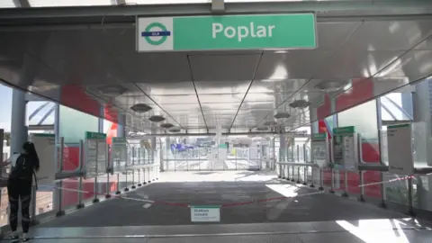 The entrance to Poplar DLR station is seen closed, with chains and a sign blocking access to the platforms, while a lone commuter stands nearby.