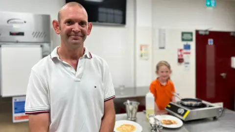 Adrian is in a white polo shirt with red and black trim smiling at the camera in a kitchen setting, with Ophelia in an orange shirt preparing food on a counter behind him. The kitchen features stainless steel appliances, white tiled walls, and an emergency exit sign above a red door.
