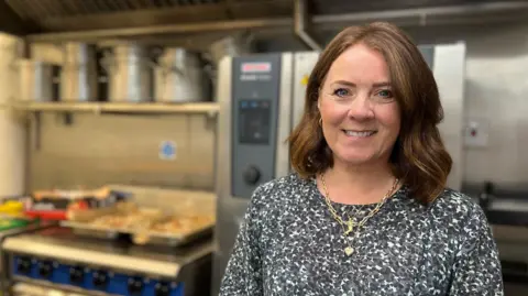 Yvonne is wearing a patterned shirt and multiple necklaces standing in a clean, professional kitchen with stainless steel appliances, including a stove with pots, an oven with blue knobs, and a ventilation hood
